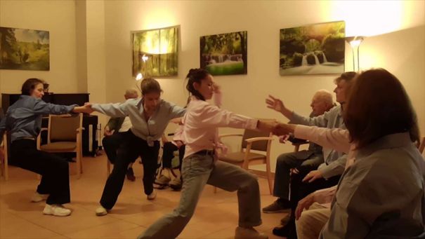 Drei Frauen führen eine Tanzsequenz in einem Raum mit gerahmten Naturfotografien an den Wänden vor, während die Bewohner zuschauen.