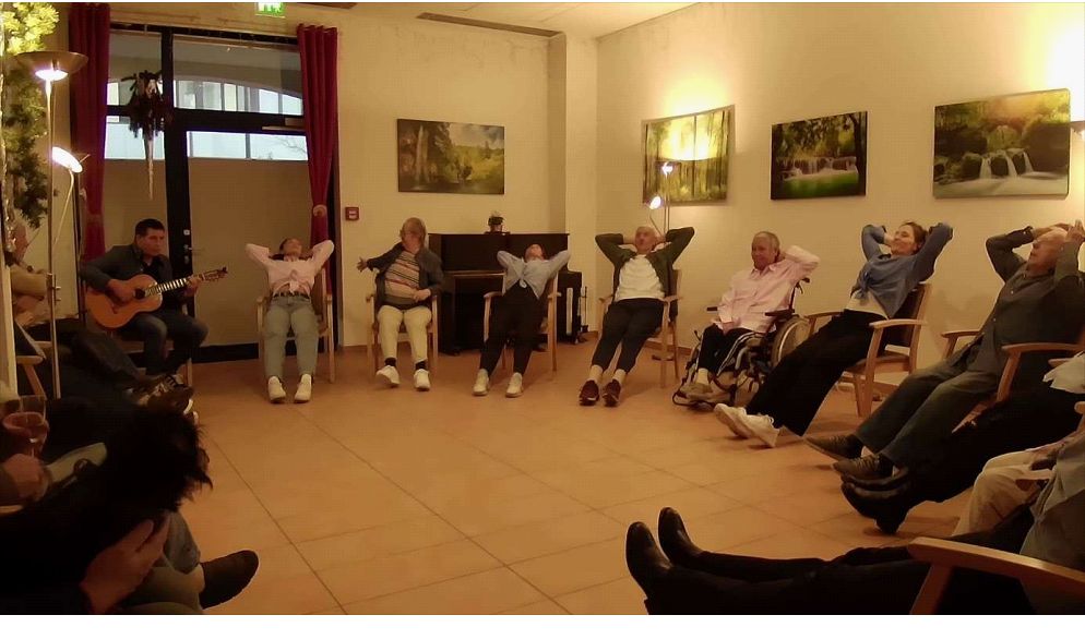 Mehrere Bewohner sitzen in einem Halbkreis in Stühlen, lehnen sich zurück und haben die Hände hinter dem Kopf, während ein Mann links Gitarre spielt.