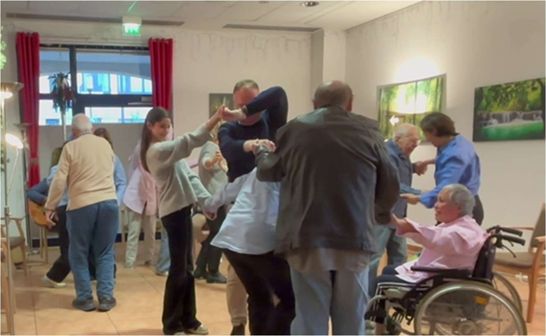 Eine Gruppe von mehreren Personen, die in einem gut beleuchteten Innenraum tanzen. Die Gruppe besteht aus jüngeren und älteren Erwachsenen, wobei einige der älteren Teilnehmer von jüngeren unterstützt werden. Eine Person sitzt rechts im Rollstuhl und hält die Hände eines anderen Teilnehmers.